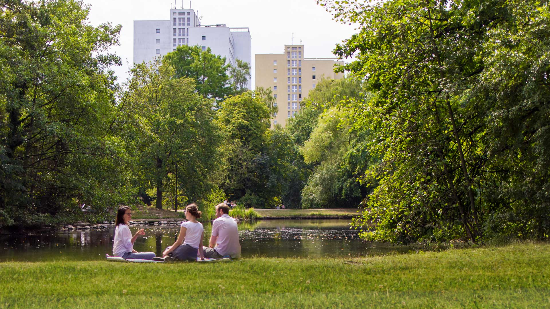 idiv-Three people sitting on grass near a pond with trees and tall buildings in the background