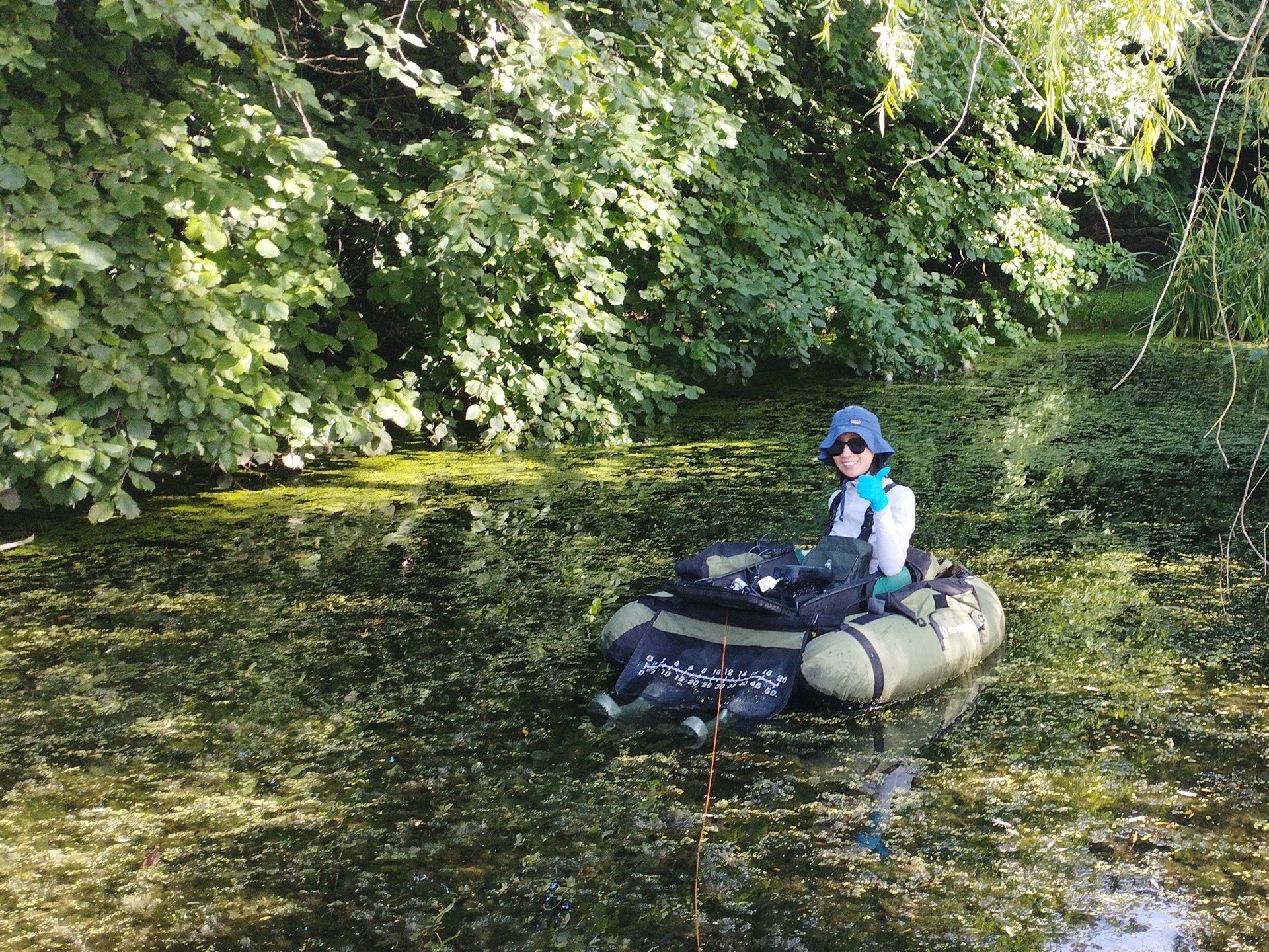 idiv-A woman with sunglasses and a large hat sits inside a small boat, afloat in a pond full of vegetation.