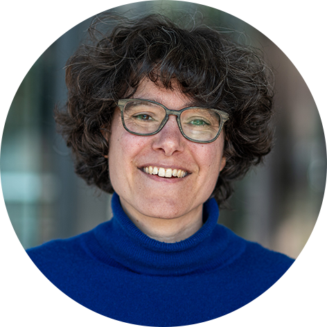 idiv-Woman (Sonja Knapp) with curly hair and glasses wearing a blue turtleneck smiling in front of a blurred background