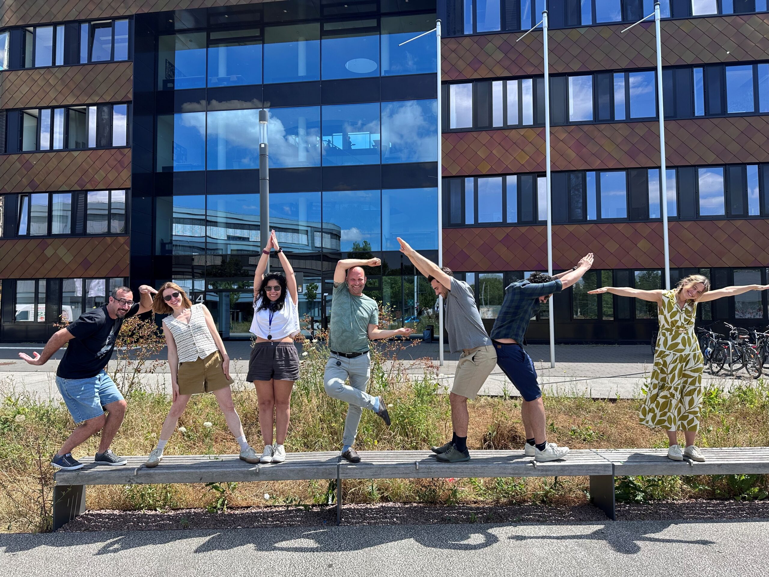idiv-Seven people standing on a bench in front of a modern building, each posing with arms and legs in varied positions forming the word